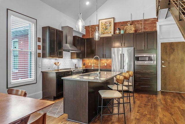 kitchen with appliances with stainless steel finishes, a sink, wall chimney range hood, dark wood-type flooring, and a breakfast bar