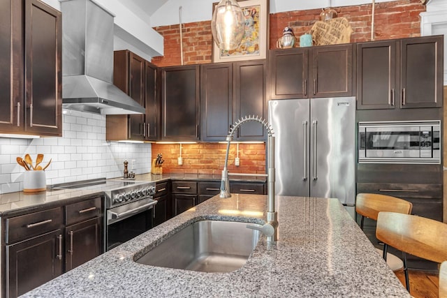 kitchen featuring premium appliances, wall chimney range hood, a sink, stone counters, and dark brown cabinets