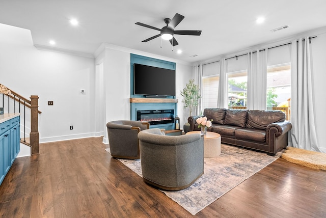living area featuring a glass covered fireplace, stairway, ceiling fan, ornamental molding, and dark wood-type flooring