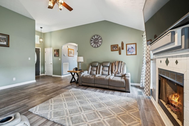 living room featuring a ceiling fan, wood finished floors, a tiled fireplace, arched walkways, and vaulted ceiling
