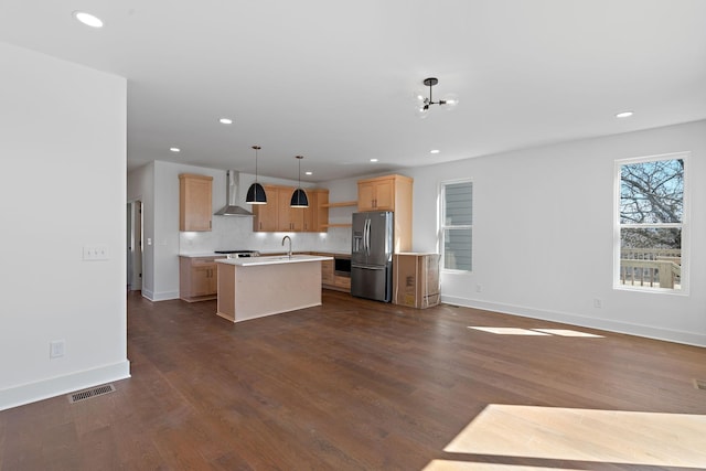 kitchen with wall chimney exhaust hood, appliances with stainless steel finishes, light brown cabinetry, light countertops, and an island with sink