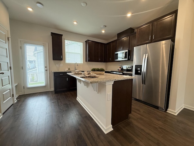 kitchen featuring stainless steel appliances, dark brown cabinetry, dark wood-style floors, a center island, and a breakfast bar