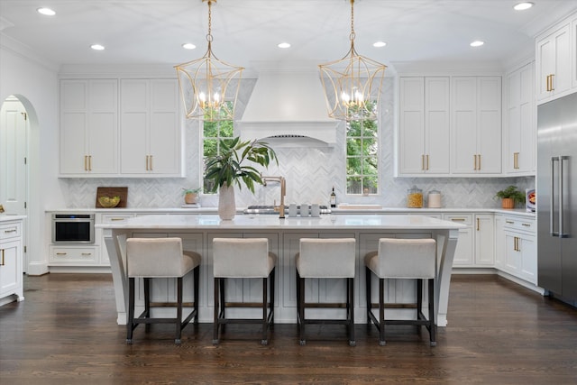 kitchen featuring custom exhaust hood, a chandelier, a kitchen island with sink, white cabinets, and backsplash