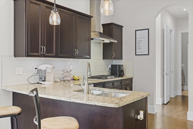 kitchen featuring wall chimney exhaust hood, dark brown cabinets, arched walkways, dark wood-style flooring, and light stone countertops