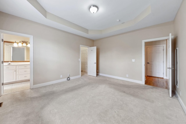 unfurnished bedroom featuring light colored carpet, a tray ceiling, baseboards, connected bathroom, and a spacious closet