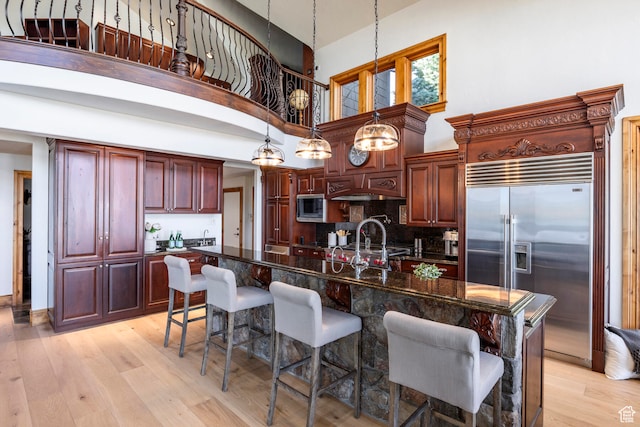 kitchen featuring built in appliances, an island with sink, decorative light fixtures, a towering ceiling, and light hardwood / wood-style floors