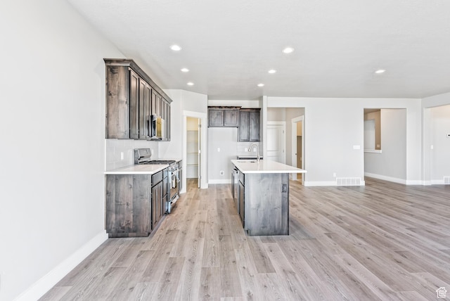 kitchen with dark brown cabinetry, stainless steel appliances, light hardwood / wood-style flooring, a kitchen island with sink, and backsplash