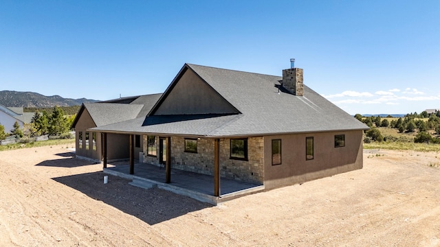 rear view of house with stone siding, a patio, a chimney, a shingled roof, and a mountain view