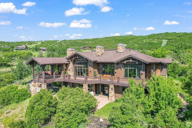 back of house featuring a forest view, stone siding, a chimney, a deck, and a patio area