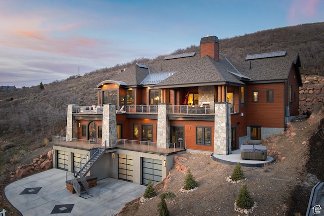 back of property at dusk with stone siding, a balcony, a chimney, a garage, and driveway