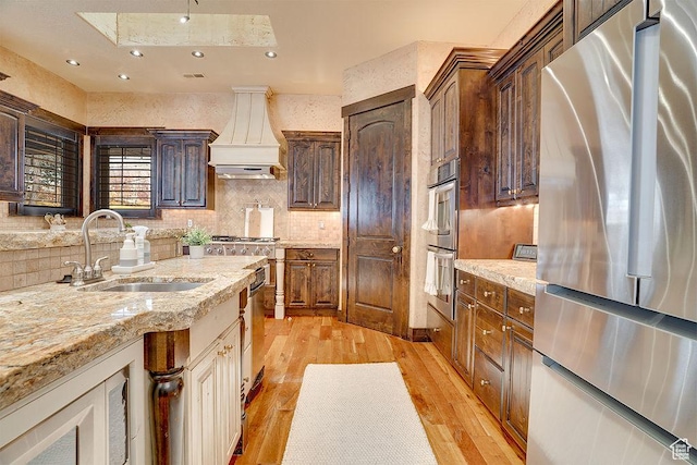 kitchen featuring a sink, appliances with stainless steel finishes, custom exhaust hood, light wood finished floors, and tasteful backsplash