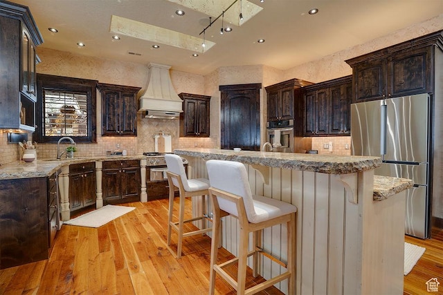 kitchen with appliances with stainless steel finishes, dark brown cabinetry, light wood-style flooring, and custom range hood