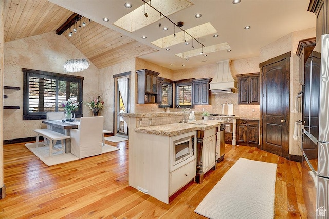 kitchen with stainless steel appliances, premium range hood, a skylight, a sink, and light wood finished floors