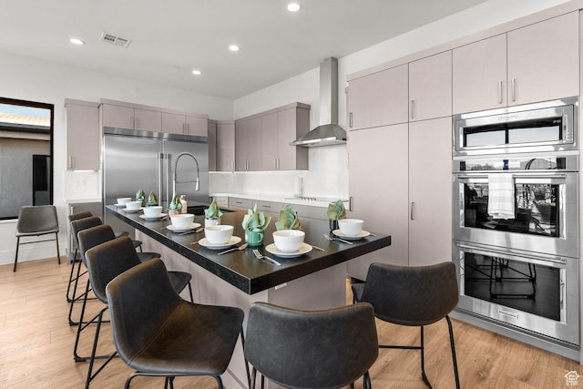 kitchen with gray cabinets, built in appliances, light hardwood / wood-style floors, a kitchen bar, and wall chimney exhaust hood