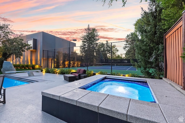 pool at dusk with a tennis court, fence, and an in ground hot tub
