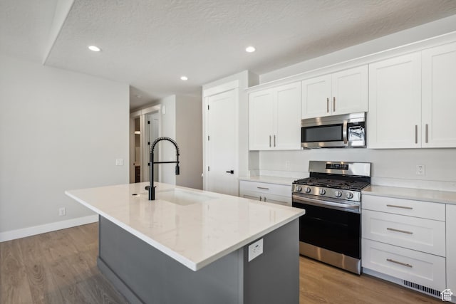 kitchen featuring a kitchen island with sink, wood finished floors, appliances with stainless steel finishes, white cabinetry, and a sink