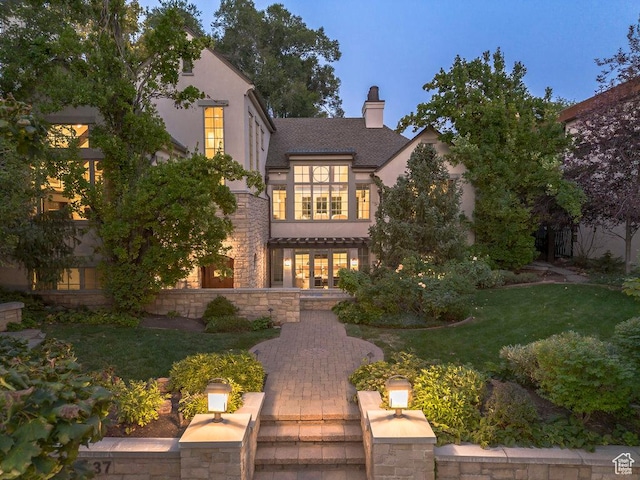 view of front of property with stucco siding, stone siding, a front lawn, a shingled roof, and a chimney