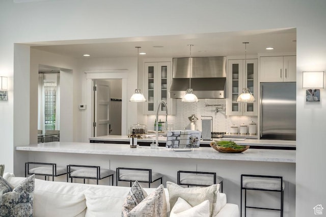 kitchen featuring white cabinets, a breakfast bar, tasteful backsplash, built in refrigerator, and wall chimney range hood