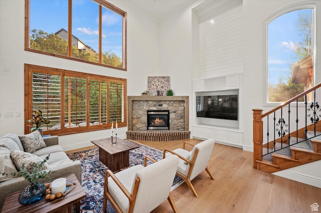 living room with crown molding, stairs, a towering ceiling, a stone fireplace, and light wood-type flooring