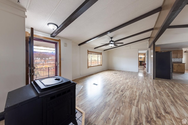 unfurnished living room featuring light wood-type flooring, a textured ceiling, a wood stove, ceiling fan, and vaulted ceiling with beams