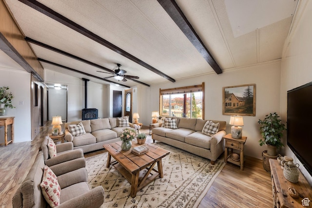 living area with light wood-style flooring, a wood stove, ceiling fan, and vaulted ceiling with beams