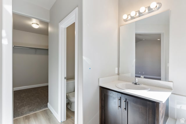 bathroom with toilet, wood finished floors, vanity, a spacious closet, and baseboards