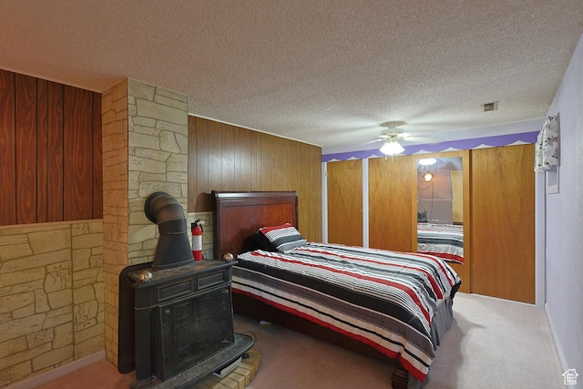 carpeted bedroom with wooden walls, a textured ceiling, a wood stove, and a ceiling fan
