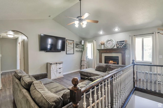 living room with a warm lit fireplace, a ceiling fan, lofted ceiling, arched walkways, and wood finished floors