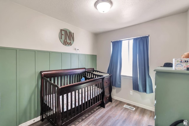 bedroom featuring visible vents, baseboards, a crib, a textured ceiling, and wood finished floors