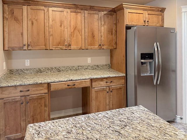 kitchen with stainless steel refrigerator with ice dispenser, brown cabinetry, built in study area, and light stone countertops