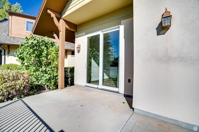 entrance to property featuring stucco siding, a shingled roof, and a patio