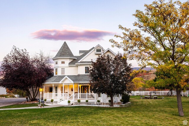 victorian house featuring covered porch, a yard, fence, and roof with shingles