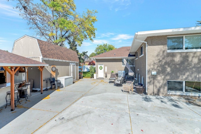 exterior space featuring a patio area, an outdoor structure, brick siding, roof with shingles, and a storage unit