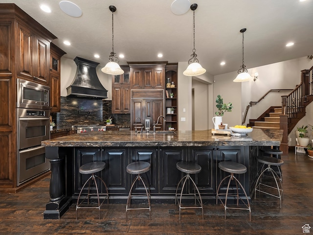 kitchen with custom exhaust hood, a large island with sink, built in appliances, and pendant lighting