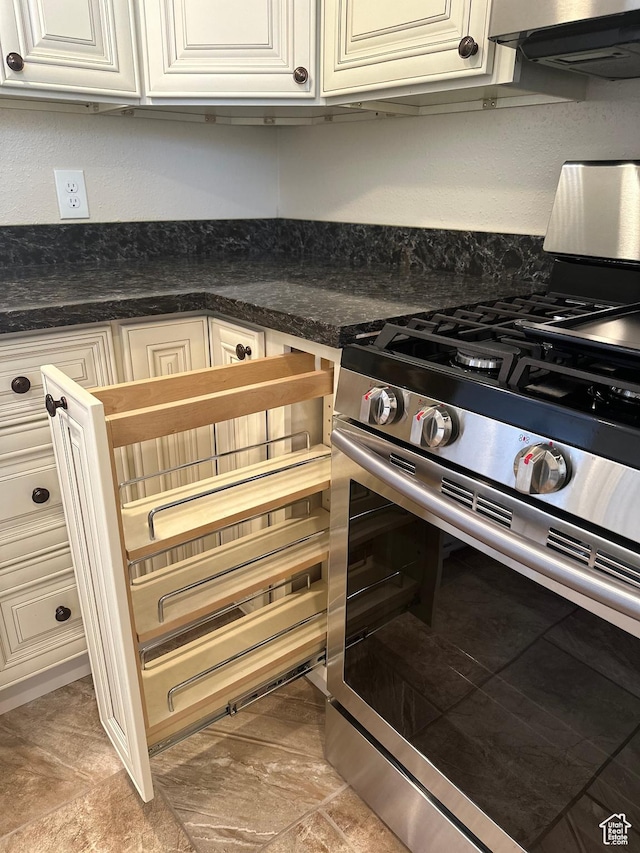 kitchen view of stainless steel range with gas stovetop, cream cabinetry, dark countertops, and range hood
