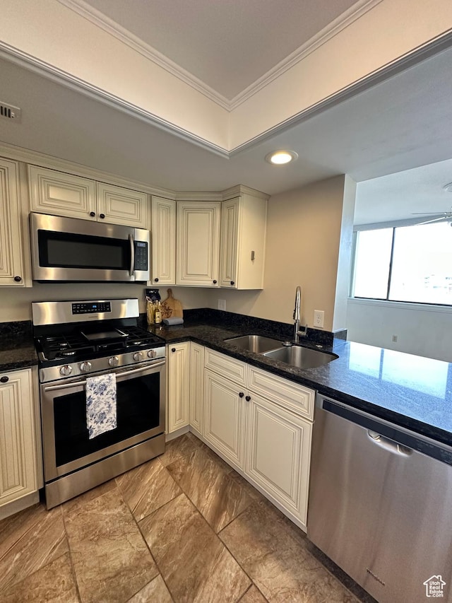 kitchen with appliances with stainless steel finishes, a sink, cream cabinets, crown molding, and dark stone countertops