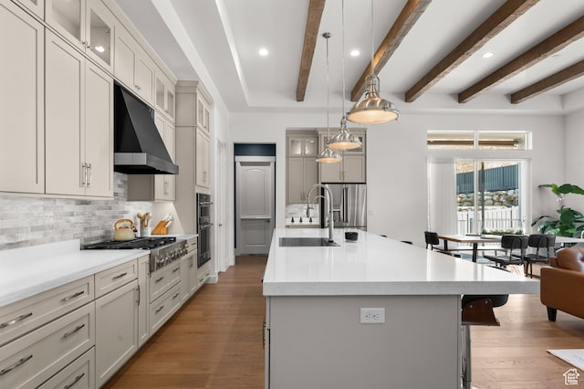 kitchen with beamed ceiling, sink, appliances with stainless steel finishes, wood-type flooring, and a kitchen island with sink