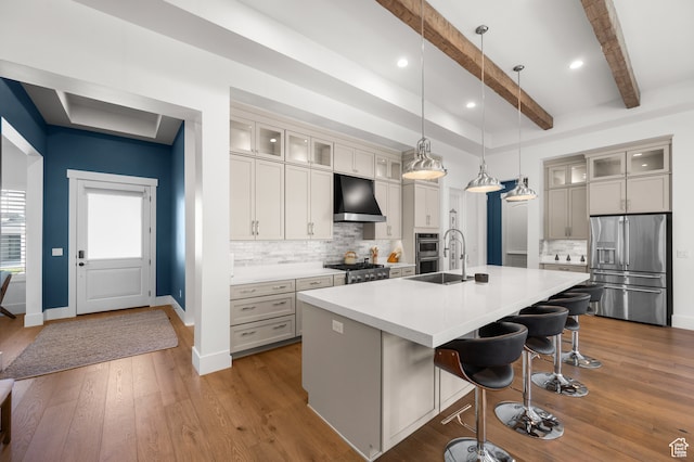 kitchen with stainless steel appliances, an island with sink, ventilation hood, beam ceiling, and decorative light fixtures
