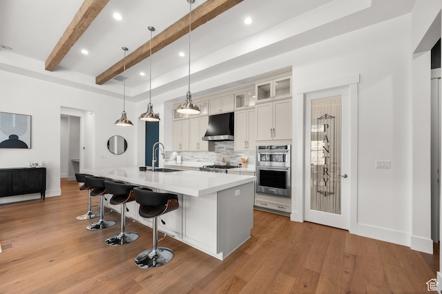 kitchen featuring ventilation hood, decorative light fixtures, beam ceiling, an island with sink, and light hardwood / wood-style flooring