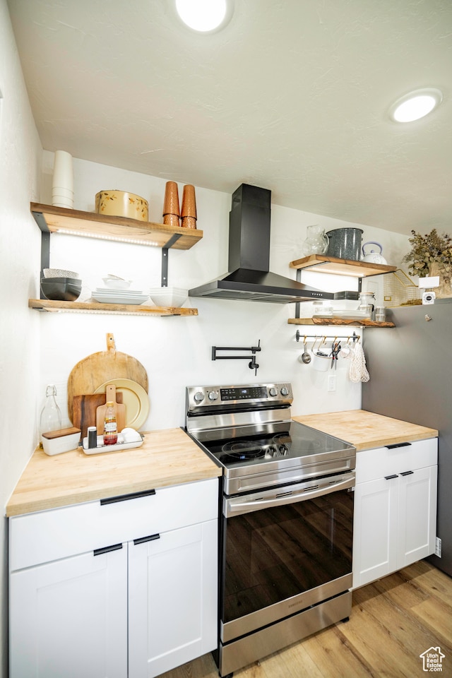 kitchen featuring butcher block counters, electric range, wall chimney range hood, white cabinets, and light wood-type flooring