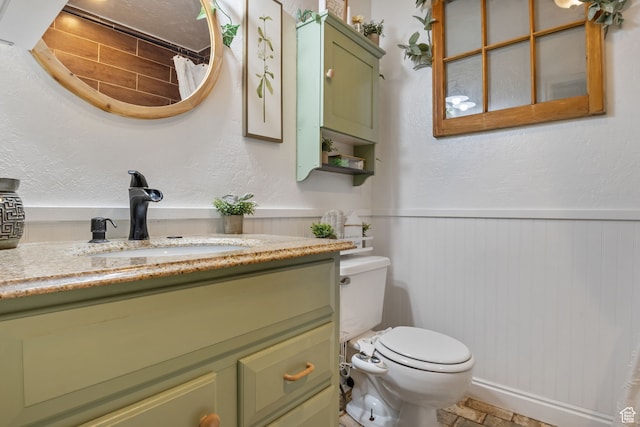 full bath featuring vanity, toilet, a textured wall, and a wainscoted wall
