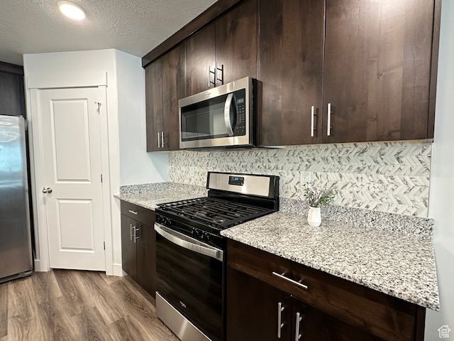 kitchen with dark brown cabinetry, tasteful backsplash, light stone counters, light hardwood / wood-style floors, and appliances with stainless steel finishes