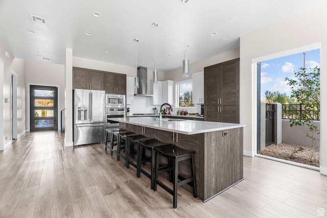 kitchen featuring dark brown cabinetry, a breakfast bar, wall chimney exhaust hood, stainless steel appliances, and a center island with sink