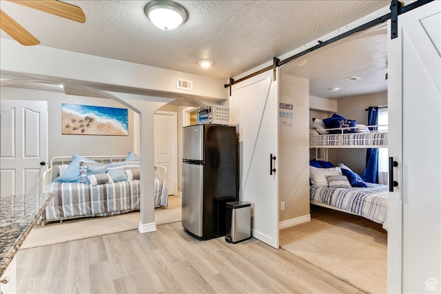 bedroom with a textured ceiling, a barn door, visible vents, freestanding refrigerator, and light wood finished floors