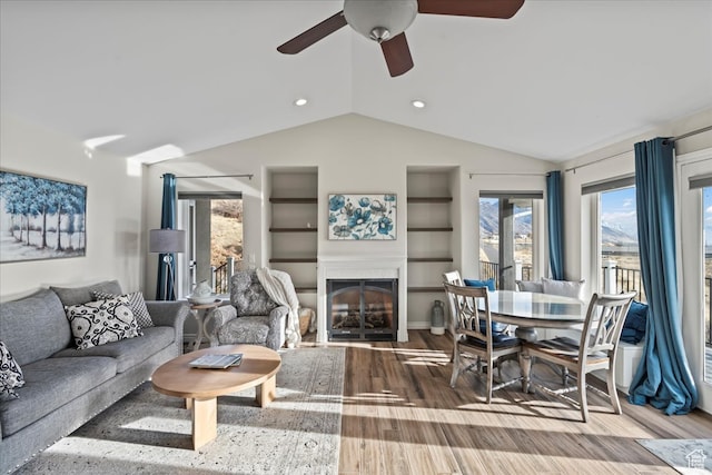 living room with built in shelves, ceiling fan, wood-type flooring, and vaulted ceiling