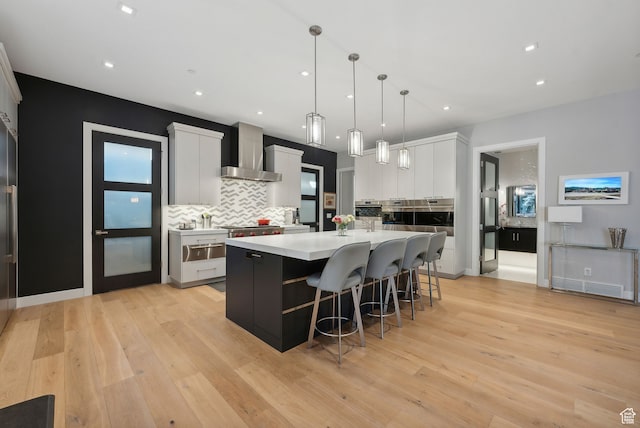 kitchen with white cabinets, wall chimney range hood, hanging light fixtures, light wood-type flooring, and a large island