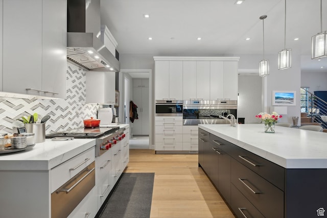 kitchen featuring wall chimney range hood, hanging light fixtures, light hardwood / wood-style flooring, an island with sink, and white cabinetry