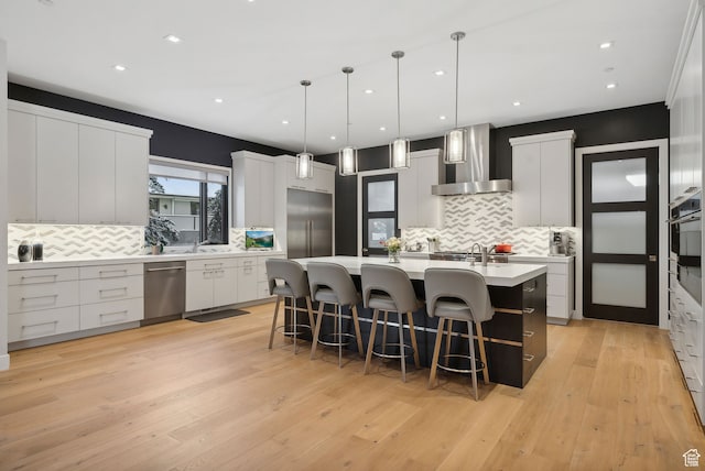 kitchen featuring wall chimney range hood, light hardwood / wood-style floors, pendant lighting, a kitchen island with sink, and appliances with stainless steel finishes