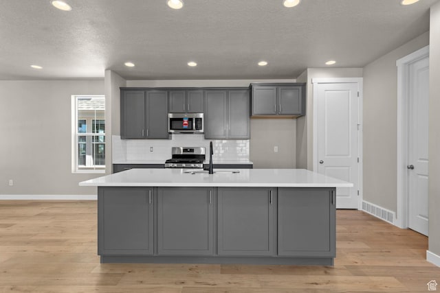 kitchen with gray cabinetry, sink, stainless steel appliances, a kitchen island with sink, and light wood-type flooring