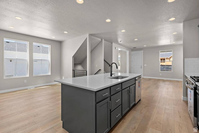 kitchen with light wood-type flooring, a textured ceiling, stainless steel appliances, sink, and an island with sink